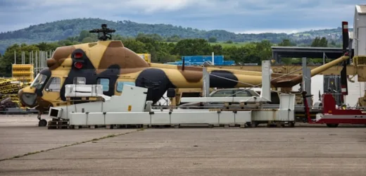 Camouflaged military helicopter on airfield, surrounded by equipment, with scenic hills and cloudy sky in the background.